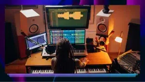 A woman with dreadlocks sits in front of several keyboards, a microphone, and a computer working on editing an audio track.