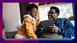 A man sits on a couch holding a tablet, he is smiling at a young boy next to him who is looking at the tablet. The young boy has a hearing aid in.