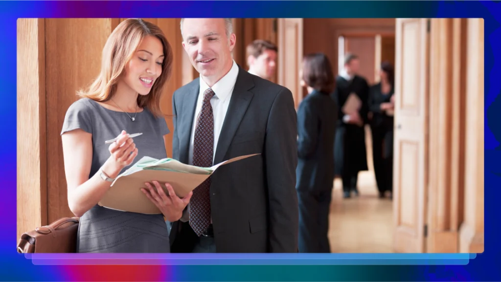 Professionally dressed man and woman discussing a file of documents outside a courtroom.