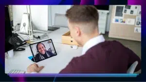A man sits facing away from the camera with his back visible. He is talking into an iPad, which is sitting on a white desk. A video with a lady smiling is on the iPad.