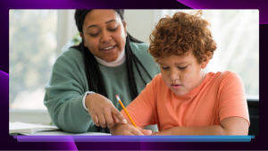 A teacher in a green shirt instructing a boy in an orange shirt, who are both sitting at a table and looking at a piece of paper. The boy is writing with a pencil.