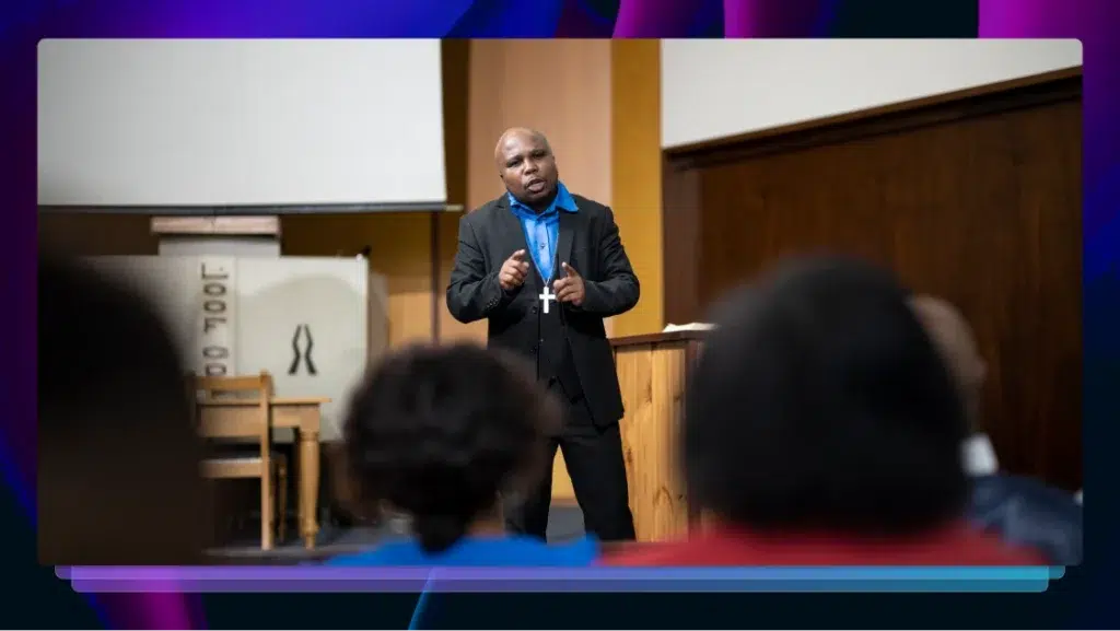 A preacher wearing a black jacket, black pants, a blue shirt, and a cross necklace preaches to his congregation.