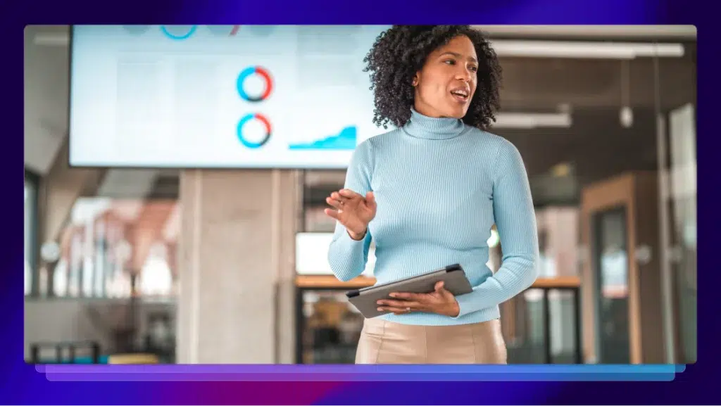 A woman in a light blue turtleneck is standing in an office. She is holding an iPad in her left hand. She is speaking to someone out of frame.