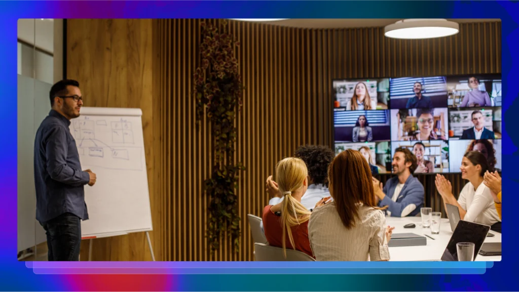 A man stands at an easel presenting a diagram to a group of his workers. Six coworkers are sitting at a table in the same room and an additional nine are on a television screen in the background virtually joining the meeting.